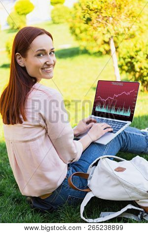 Smiling Attractive Woman Using Laptop With Traiding Appliance In Park And Looking At Camera