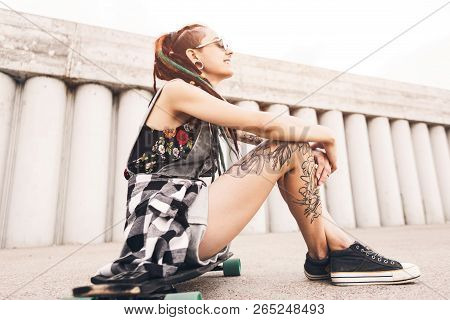 Young Girl With Tattoo And Dreadlocks Sitting On A Longboard On The Background Of Concrete Structure