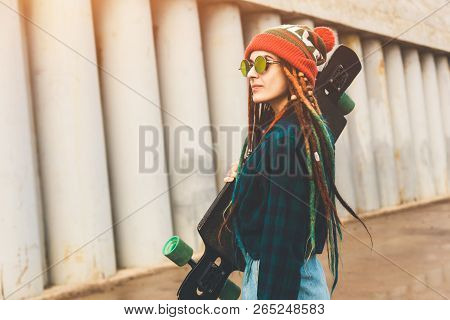 Young Girl In Sun Glasses And A Cap Stands With A Longboard Against The Background Of Concrete Struc