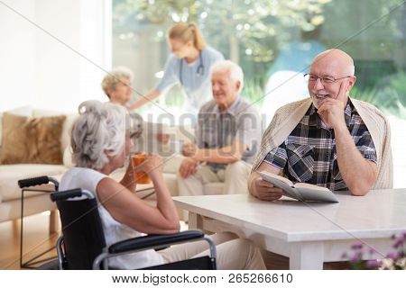 Elderly Woman In The Wheelchair Drinking Tea During Conversation With Smiling Friend