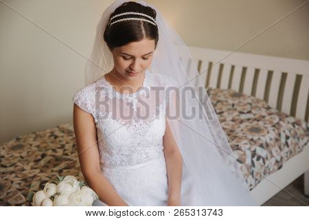 Beautiful And Tenderness Bride In White Dress And Veil. She Is Sittind On The Bed