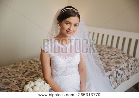 Beautiful And Tenderness Bride In White Dress And Veil. She Is Sittind On The Bed