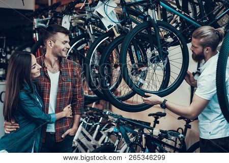 Consultant Helps Young Couple In Bike Choosing. Bearded Shop Assistant Shows Wheel To Beautiful Girl