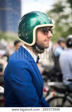 Causasian Man In Blue Suit With Bowtie, Sunglasses And Motorbike Helmet With Crowd In Background.