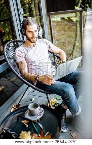 Poortrait Of A Handsome Man Working With Laptop And Headphones On The Balcony Of The House In The Be