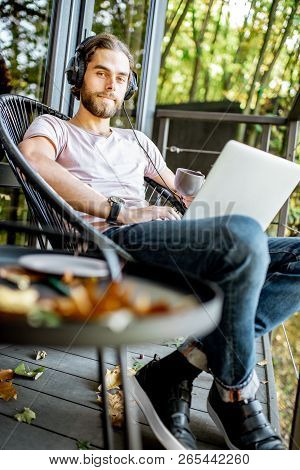 Poortrait Of A Handsome Man Working With Laptop And Headphones On The Balcony Of The House In The Be