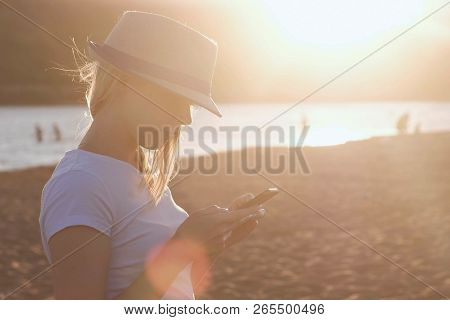 Beautiful Blonde Girl In A Hat Types A Message On Her Mobile Phone On The Beach At Sunset.