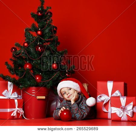 Funny Smiling Childboy In Santa Red Hat Lying On  Christmas Tree  Holds In One Hand A Christmas Ball