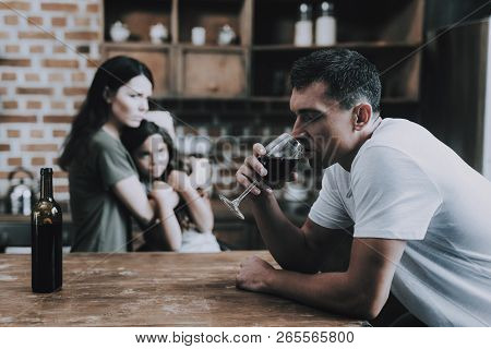 Mother And Child Look At Drunk Father In Kitchen. Man Sits On Chair In Modern Room In Wooden Style H