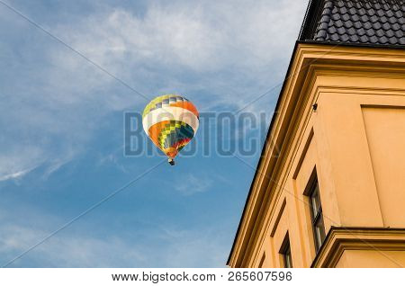 Colorful Color Hot Air Balloon In Blue Sky With White Clouds Over Roofs Of Traditional Typical Yello