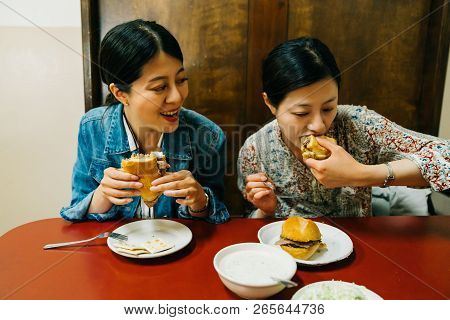 Hungry Ladies Sitting In The Red Table And Joyfully Eating Hamburgers. Asian Sisters Having Lunch At