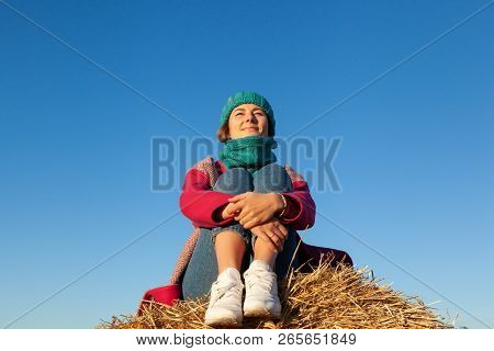 Young Attractive Lady In Blue Knitting Hat And Pink Coat Sitting On Haystack. Smiling Beautiful Hips