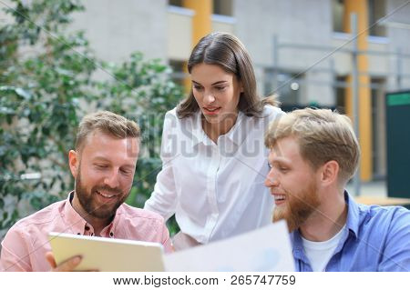 Group Of Young People In Casual Wear Sitting At The Office Desk And Discussing Something While Looki