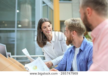 Group Of Young People In Casual Wear Sitting At The Office Desk And Discussing Something While Looki