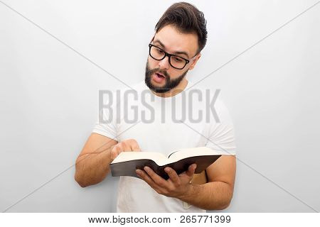 Picture Of Young Man Reading Aloud From Book. He Holds It In Hands And Points On It. Young Man Is Co