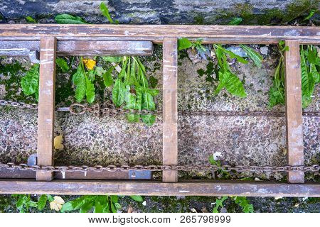 An Old Wooden Ladder Stepladder With Rusty Large Chains Lies On A Cement Surface Interspersed With S