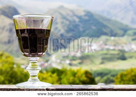 A Glass Of Georgian Red Wine Stands On A Wooden Railing On The Background Of The Georgian Village An