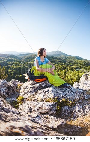 A Woman Is Sitting In A Sleeping Bag At Sunset. Camping On Top Of A Mountain. A Smiling Girl Rests A