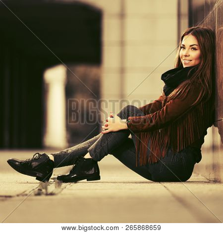 Happy young woman sitting at the wall in city street  Stylish fashion model wearing brown leather fringe suede jacket and dark blue jeans