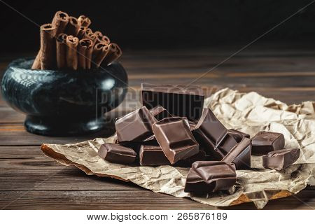 Chunks Of Broken Chocolate On Baking Paper And Cinnamon Sticks In Marble Bowl On A Wooden Table. Dar