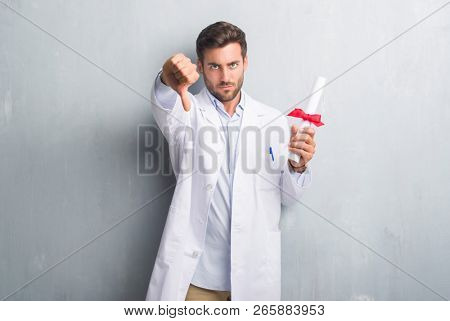 Handsome young doctor man over grey grunge wall holding diploma with angry face, negative sign showing dislike with thumbs down, rejection concept