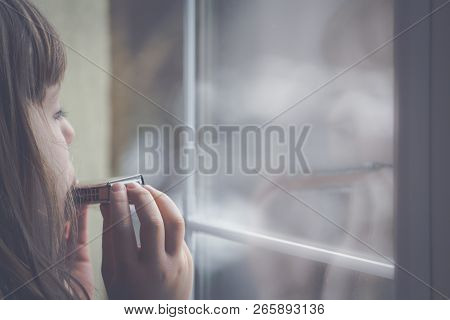 Sad Caucasian Girl Standing By The Window And Playing Harmonica At Home