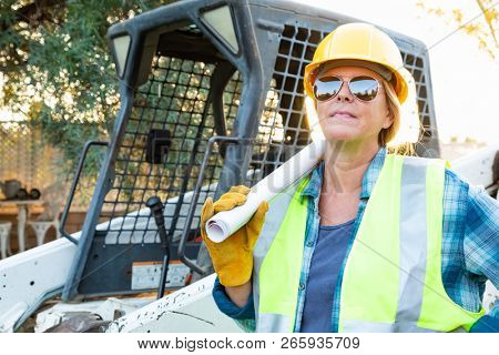 Female Worker Holding Technical Blueprints Near Small Bulldozer At Constrcution Site