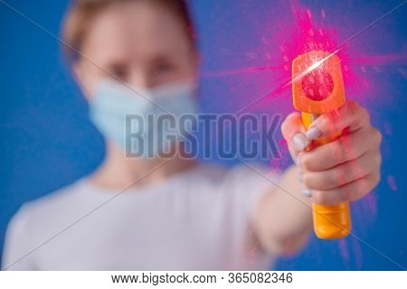 Woman In Medical Face Mask Holding Yellow Pyrometer To Measure Temperature Toward Camera - Close Up 
