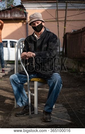 Senior Man In Protective Mask With A Cane Sitting On A Chair In The Garden In Sunny Day