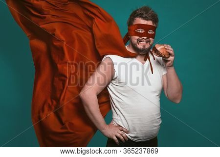 Hungry Super Hero Man Eating A Hamburger, Super Hero Or Antihero In A Red Suit With A Flying Cloak E