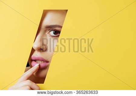 Woman Looking At Camera Across Quadrangular Hole In Yellow Paper And Touching Pink Lip
