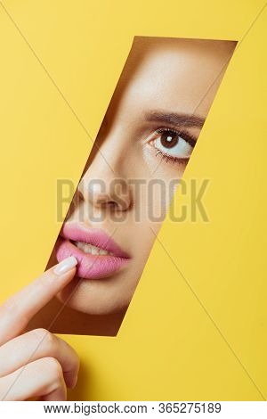 Woman Looking Across Quadrangular Hole In Yellow Paper And Touching Pink Lip