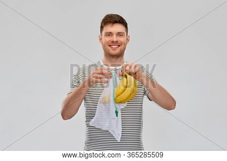 food, sustainability and eco living concept - smiling young man in striped t-shirt putting bananas into reusable string bag over grey background