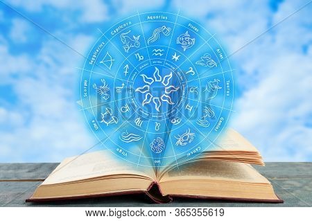 Old Book On Wooden Table And Illustration Of Zodiac Wheel With Astrological Signs Against Blue Sky
