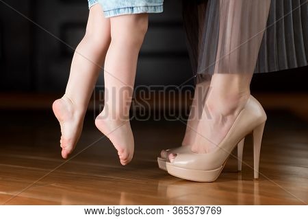 A Little Girl With Bare Feet Reaches For A Shod Mom In The Hallway At Home. Mom Takes The Baby And T