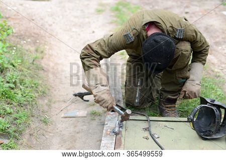 Old Man Welder In Brown Uniform Prepares Metal Door Surface For Welding With Arc Welding Machine Out