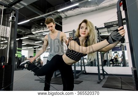 Sporty Girl Doing Stratching Legs Exercises With Assistance Of Her Personal Trainer At Public Gym. C