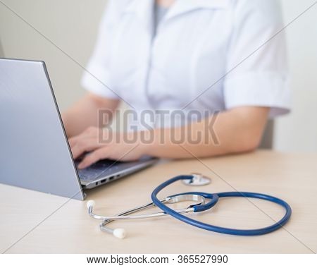 Unrecognizable Female Doctor In Uniform Uses A Laptop. Faceless Medical Worker Writes A Report On Th