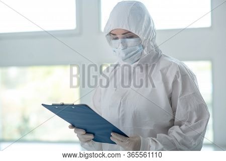 Medical Worker In Protective Clothing, Medical Mask And Goggles Checking Something On Clip Folder