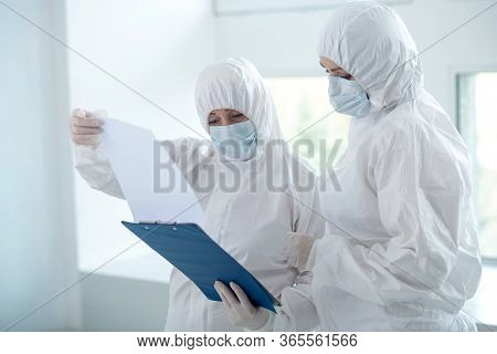 Medical Workers In Protective Clothing And Medical Masks Looking Through Papers On Clip Folder