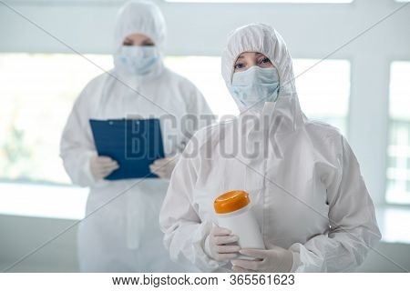 Medical Worker In Protective Clothing Holding Disinfecting Wipes Bottle, Her Colleague Standing Behi