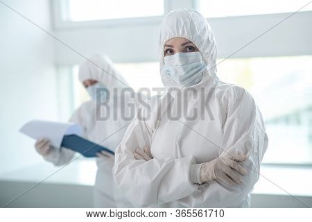 Female Medical Worker In Protective Clothing Standing With Folded Arms, Her Colleague Standing Behin