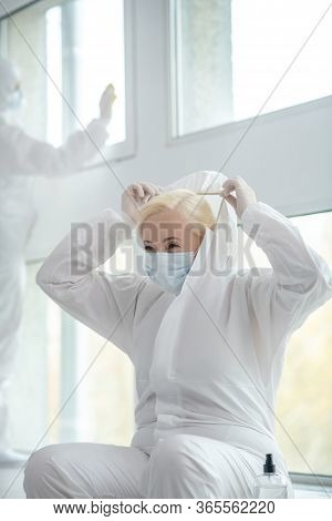 Blonde Medical Worker In Protective Clothing And Medical Mask Sitting Next To The Window, Putting On