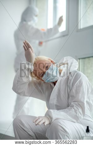 Tired Blonde Medical Worker In Protective Clothing And Medical Mask Sitting Next To The Window,