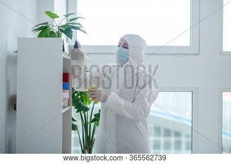 Medical Worker In Protective Clothing And Medical Mask Sprays Disinfectant On Closet