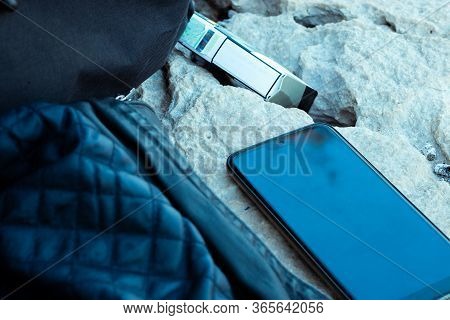 Smartphone Laying On A Rock With A Pack Of Cigarettes Behind It. Jacket And Backpack Next To It, Ear