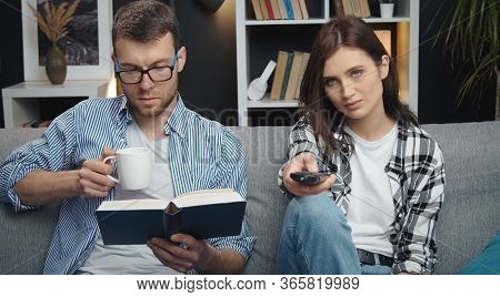 Spouses Trying To Divert Themselves Sitting On Sofa, Woman Changing Tv Channels While Man Reading Bo