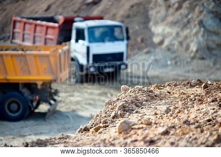 Pile Of Ore Rocks Over Out Of Focus Background With Two Heavy Multi-ton Heavy Mining Trucks Exportin