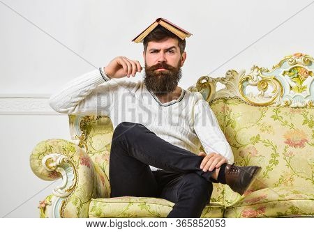 Macho Sits With Open Book On Head, Like Roof. Man With Beard And Mustache Sits On Baroque Style Sofa