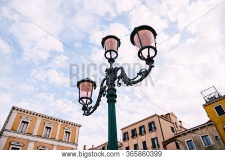 An Ancient Triple Metal Street Lamp, With Pink Plafonds Against The Sky And Buildings Of Venice, Ita